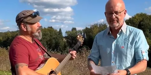 Ein Mann mit Gitarre und ein anderer Mann mit Textblatt 